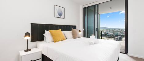 A modern bedroom with a large bed, a nightstand with a lamp, a city view through a sliding door, and a framed artwork on the wall.