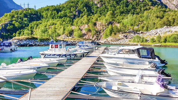 Un puerto deportivo con barcos amarrados a lo largo de un muelle de madera, rodeado por un paisaje montañoso con abundante vegetación.