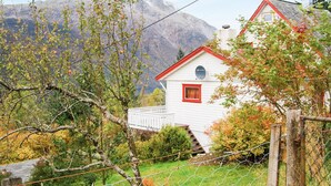Una casa blanca con un ventanal rojo y una antena parabólica, rodeada de árboles y montañas.