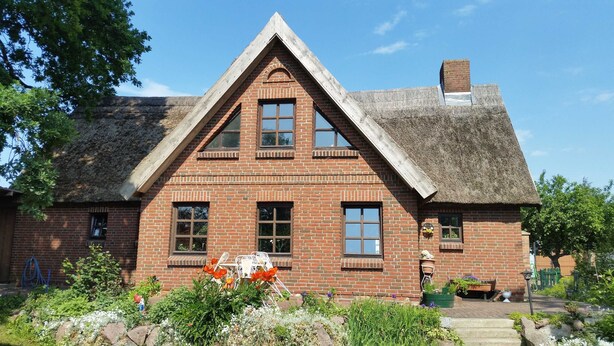 A traditional brick house with a steeply pitched thatched roof, multiple windows, and a well-maintained garden with flowers and plants. - Holiday house Rankwitz - holiday house (Rankwitz)