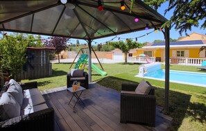 Un patio techado al aire libre con mobiliario de mimbre, una mesa con bebidas y un conjunto de columpios al fondo.
