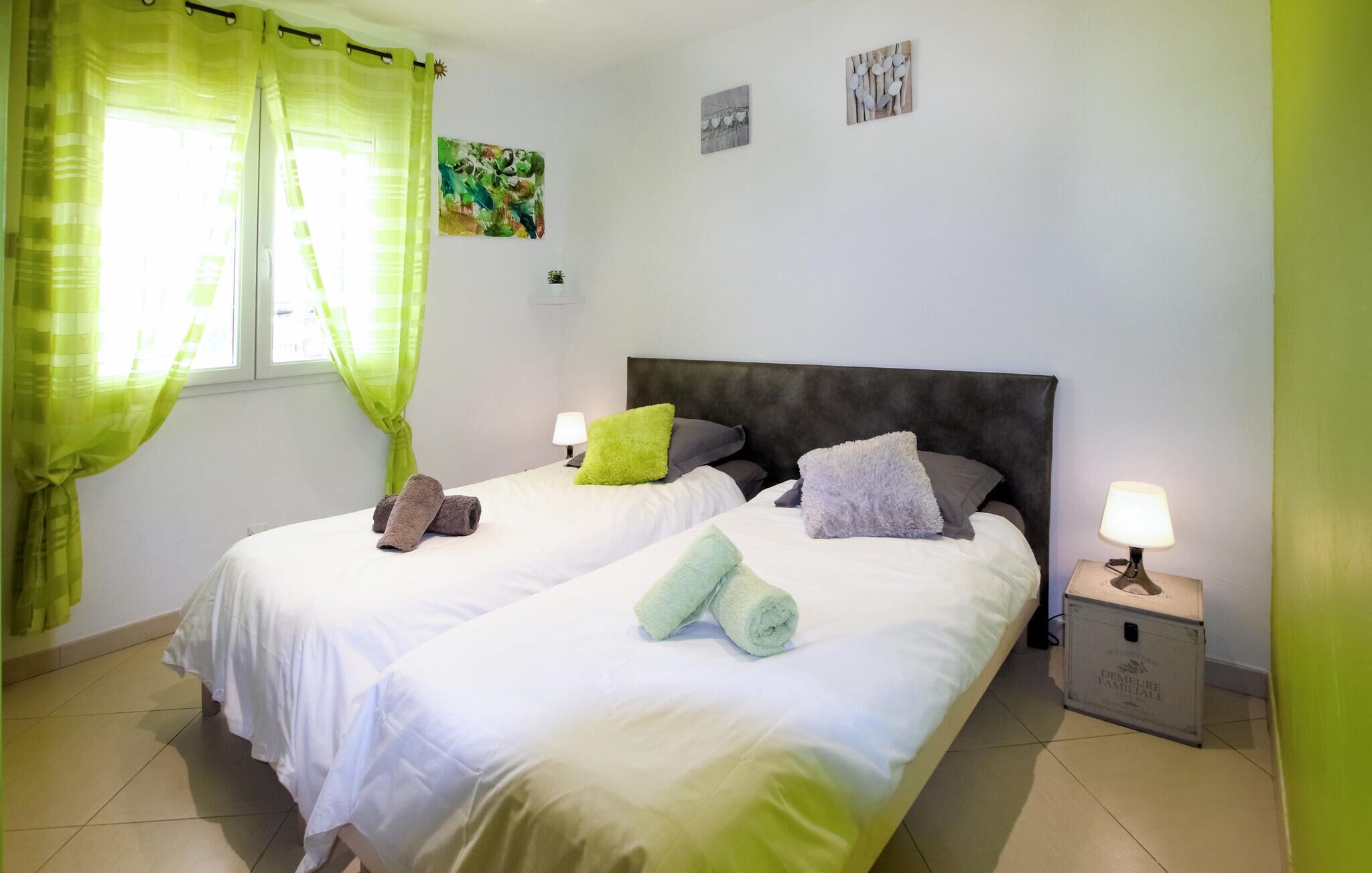 A hotel room with two beds, green curtains, a nightstand with a lamp, and artwork on the walls.