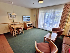 Ein Hotelzimmer mit Esstisch, Stühlen, einer Couch, einem Fernseher und einem Fenster mit Vorhängen.