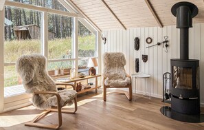 A cozy room with two fur-covered chairs, a wooden table, a fireplace, and a large window overlooking a forest. - Lovely home in Rømø with sauna (Rømø)