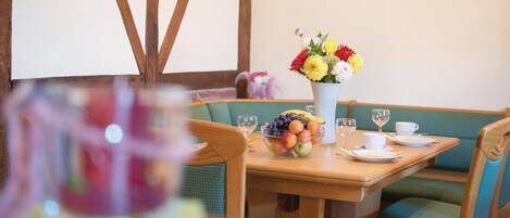Comedor con mesa de madera, un cuenco de fruta, un jarrón de flores y asientos acolchados verdes.