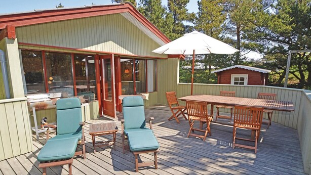 Eine Holzterrasse mit Tisch und Stühlen, ein überdachter Bereich mit einer Tür und ein kleines Gartenhaus im Hintergrund.
