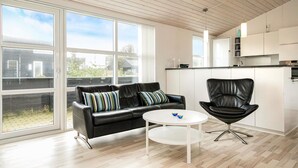 Un salon moderne avec un canapé en cuir noir, une table basse blanche et un fauteuil en cuir noir. De grandes fenêtres offrent une vue sur l’extérieur.