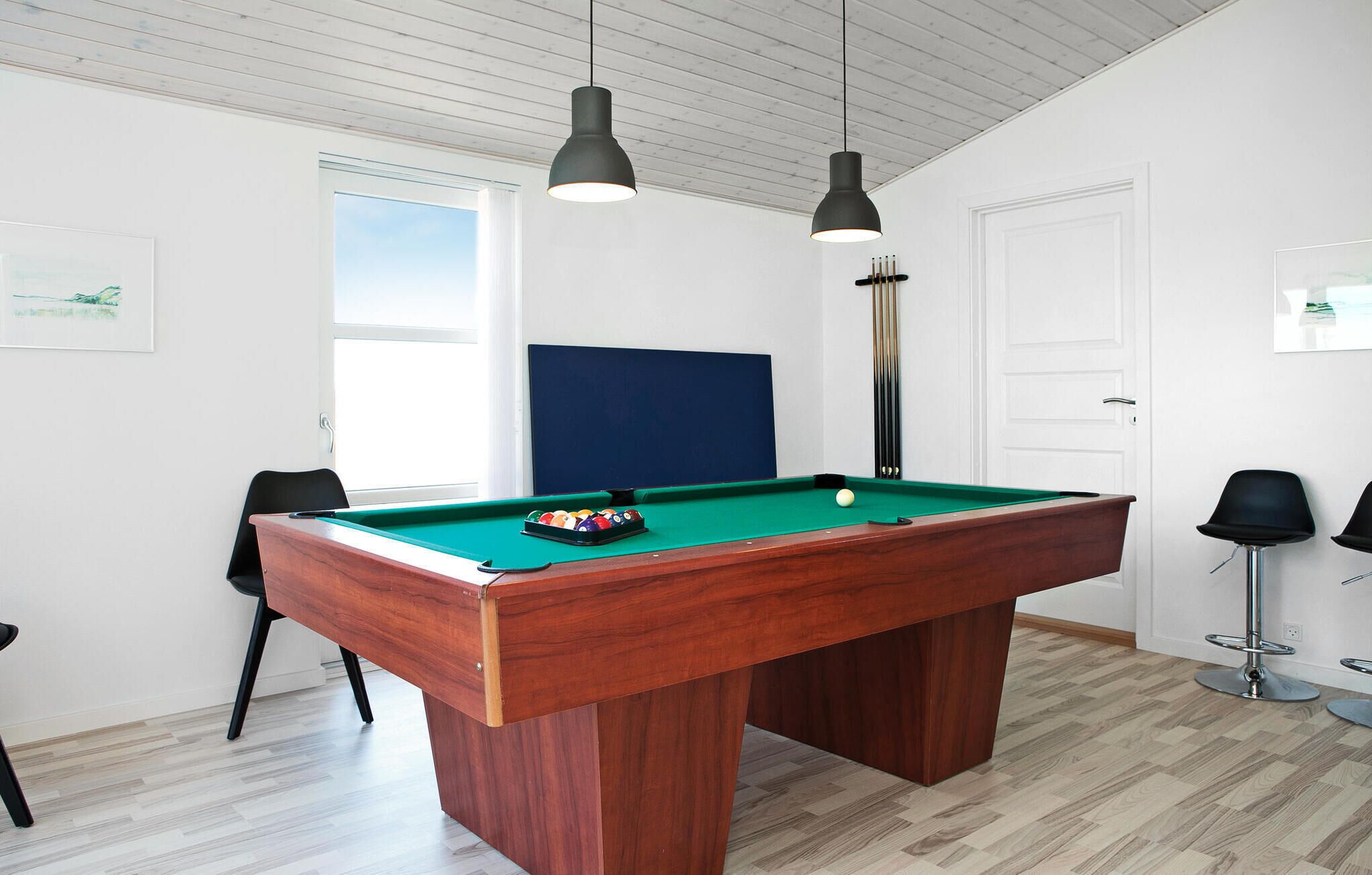 Une table de billard avec un cadre en bois, deux lampes suspendues, une tête de lit rembourrée bleue et une porte dans une pièce dotée d’un plancher en bois et de murs blancs.