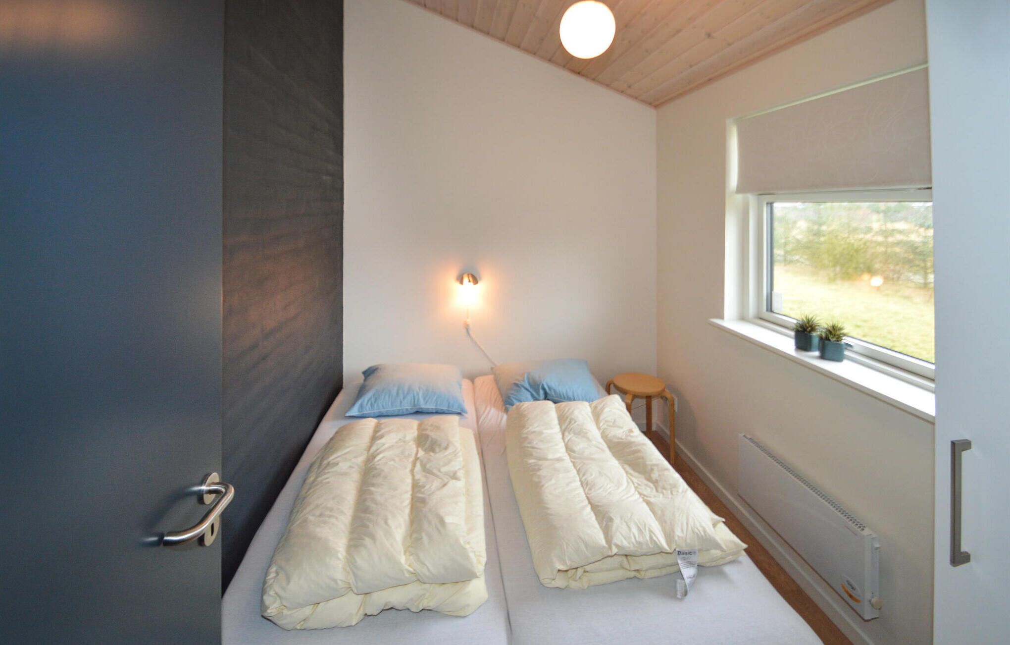Une petite chambre compacte, équipée de deux lits simples, d’une table de chevet, d’une lampe fixée au mur et d’une fenêtre donnant sur un paysage verdoyant.