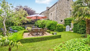 Ein Steinhaus mit einer Terrasse, auf der sich ein Tisch und Stühle unter einem Sonnenschirm befinden, umgeben von üppigem Grün und einem gepflegten Rasen.