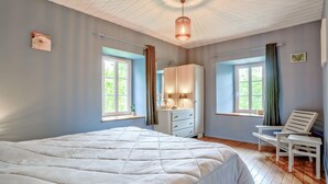 Ein Schlafzimmer mit einem großen Bett, zwei Fenstern mit Vorhängen, einem Nachttisch, einem Sessel und einem Kleiderschrank.