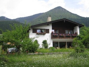 Un bâtiment à deux étages doté d’un balcon, entouré de verdure et avec des montagnes en arrière-plan.