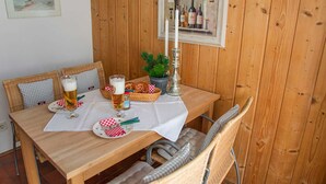 Puuinen ruokapöytä, jonka päällä on valkoinen pöytäliina, ja pöydän ympärillä on punottuja tuoleja. Seinällä on kehystetty kuva.