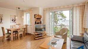 Un salon comprenant une table à manger en bois, des chaises, un canapé, un téléviseur et une vue sur la mer grâce à de grandes fenêtres.