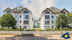 Un immeuble résidentiel moderne à deux étages, doté d’une façade symétrique, de grandes fenêtres et d’une clôture blanche en bois devant l’entrée.