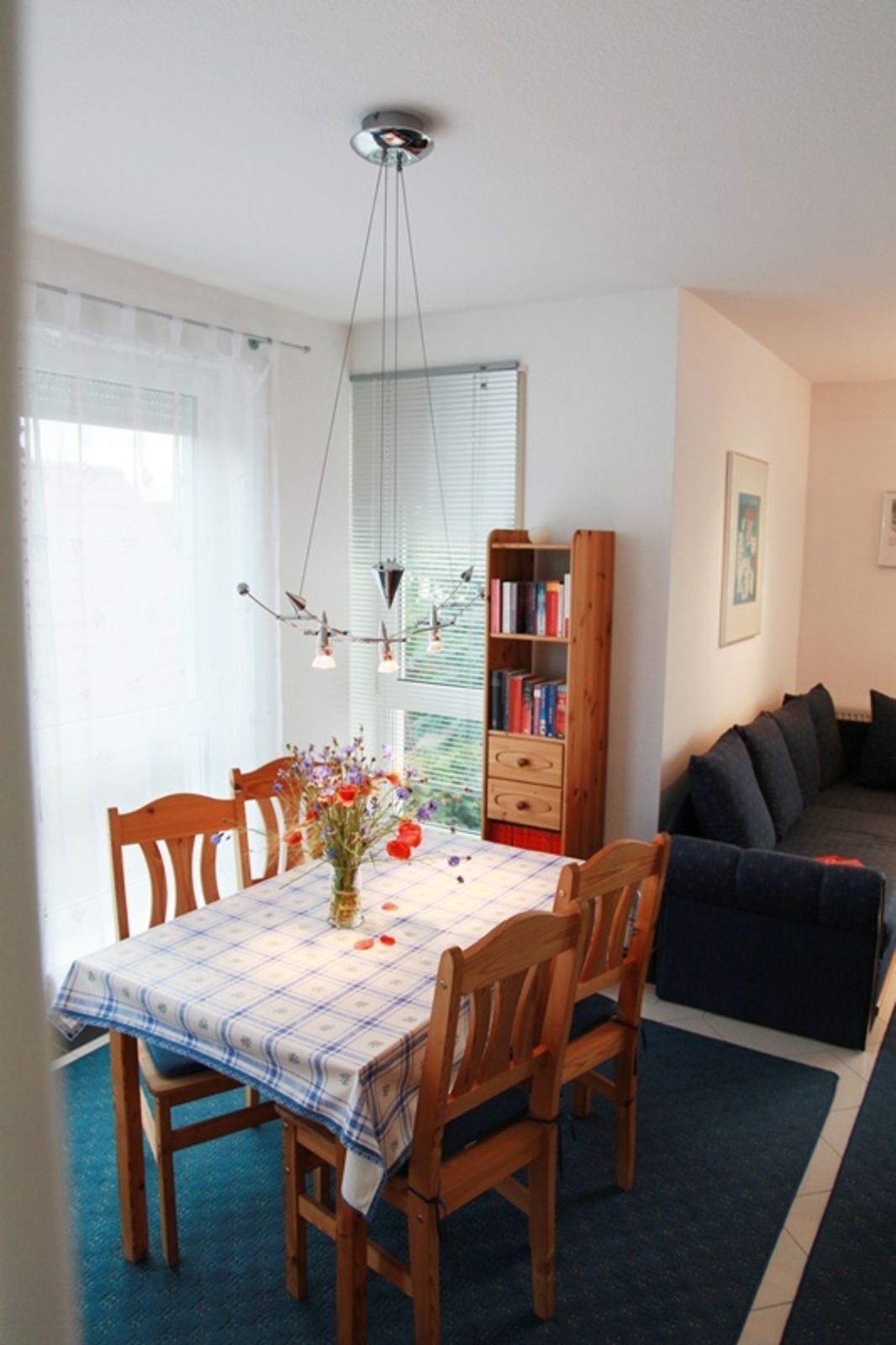 Un coin repas avec une table dressée pour quatre personnes, entourée de chaises en bois. Une bibliothèque remplie de livres est adossée au mur, et un canapé de couleur sombre est visible au fond.