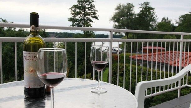 A table with a bottle of wine and two glasses on a balcony overlooking a garden and a red-roofed building. - Vacation apartment on Lake Mirower See (with balcony) left Whg.2 - Ferienwohnungen am See (Mirow)
