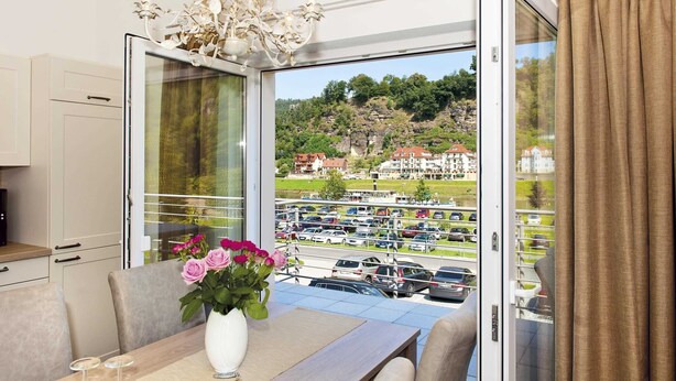 Ein Zimmer mit Esstisch, Blumen in einer Vase, einer Chandelierleuchtre und Blick auf einen Parkplatz und Gebäude durch eine Glastür.