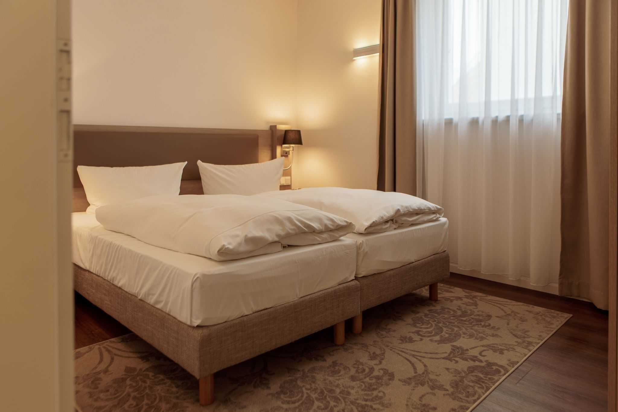Ein Doppelbett mit weißer Bettwäsche, eine Nachttischlampe, ein gemustertes Teppich und durchscheinende Vorhänge.