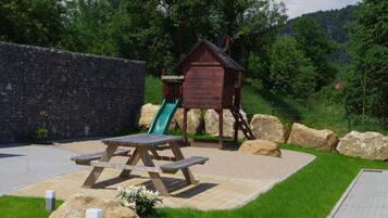 A picnic area with a wooden bench and a slide, surrounded by a stone wall and greenery.