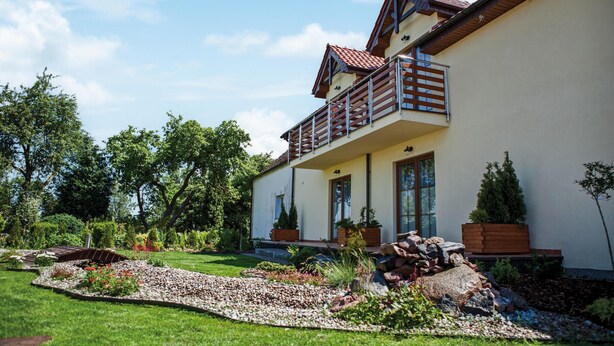 A two-story house with a balcony, surrounded by a well-maintained garden with various plants and a pebble pathway.
