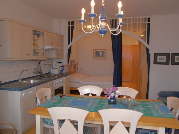 A compact kitchen with a dining area, including a table with chairs, a chandelier, and a bed in the background.