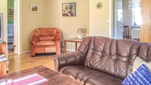 Un salon avec un canapé en cuir marron, une table basse en bois et une petite table d’appoint avec une lampe. Un tableau encadré est accroché au mur, et une porte donne accès à une autre pièce.