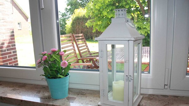 Ein Fenster mit einem türkisfarbenen Topf voller pinkfarbener Blumen und einem weißen Laternen mit Kerze darin.