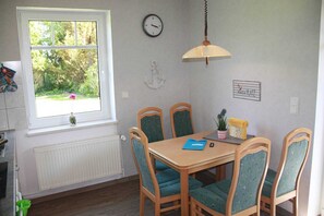 Un coin repas avec une table et des chaises, une fenêtre donnant sur un espace verdoyant, une horloge murale et une enseigne indiquant « Kleiner Platz ».