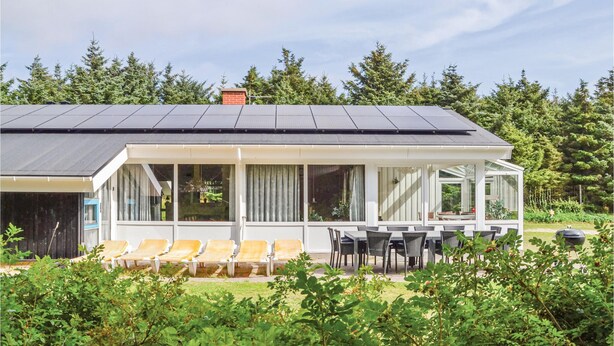 Une maison moderne avec un toit équipé de panneaux solaires, un patio couvert avec des meubles d’extérieur, et une forêt verdoyante en arrière-plan.