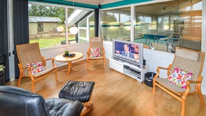 Un salon meublé de meubles en bois, avec un téléviseur et de grandes fenêtres donnant sur un jardin.