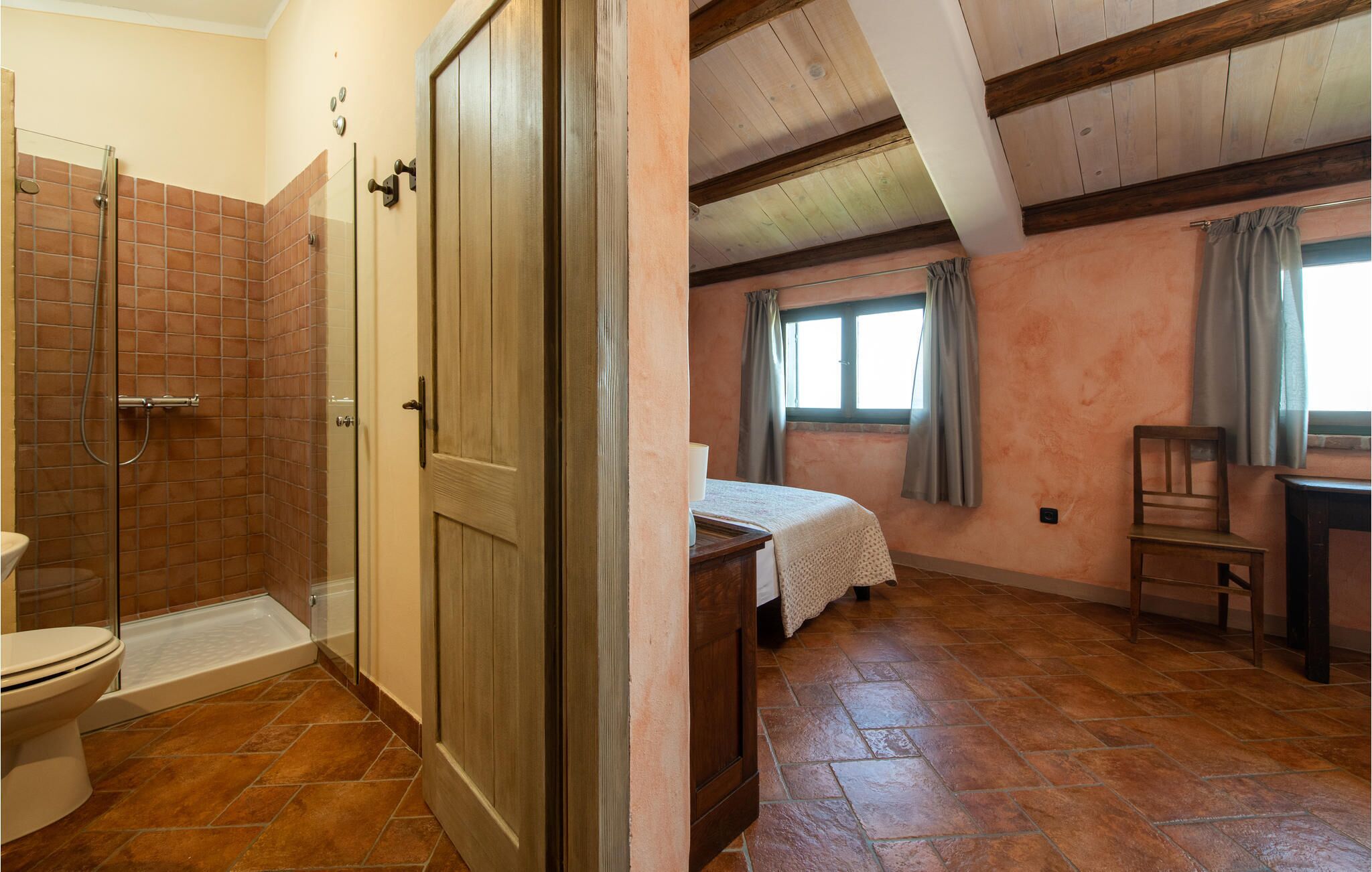 A bedroom with a wooden door, a bed with a white bedspread, a window with curtains, and a wooden chair.