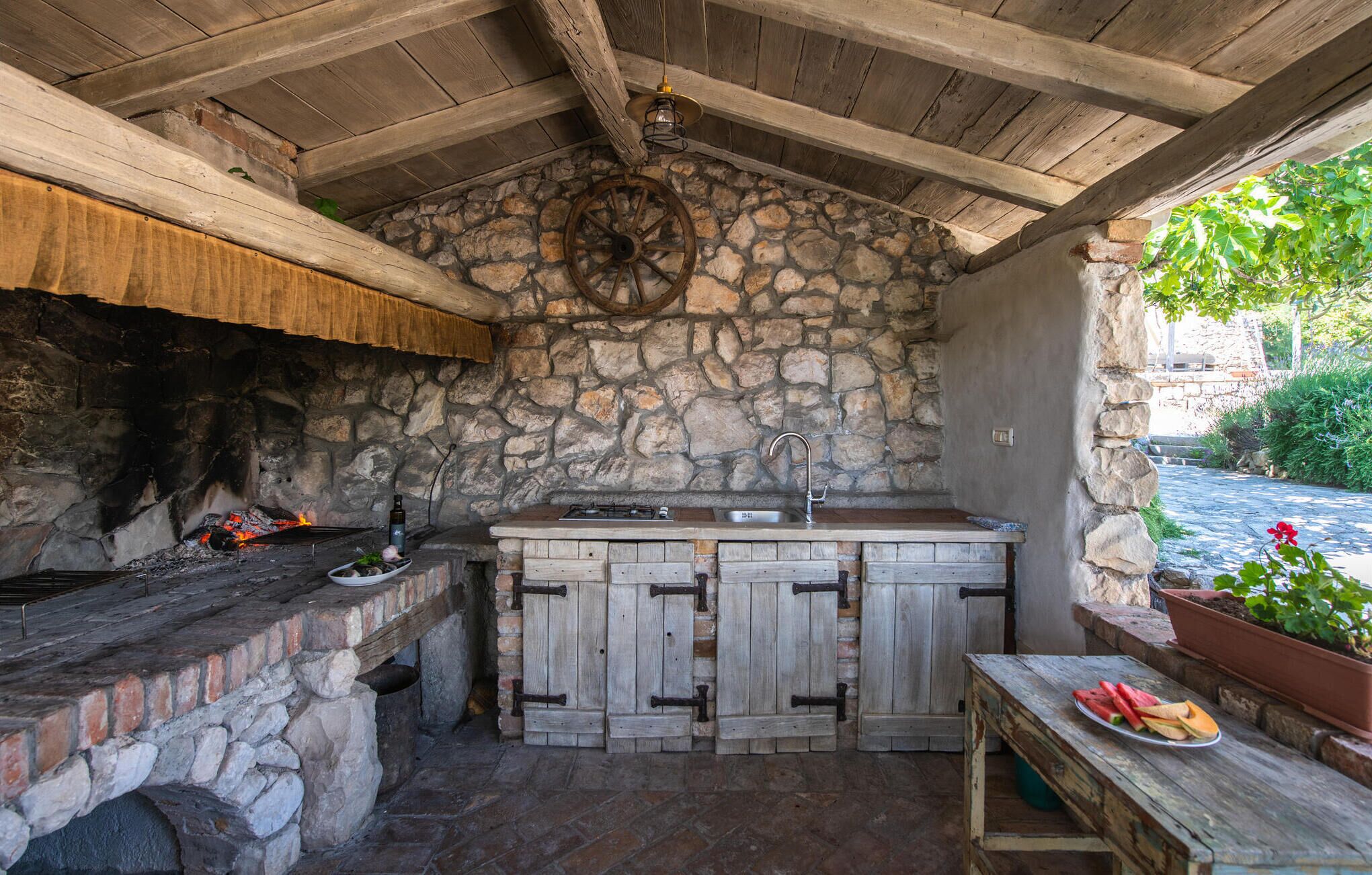 Cocina exterior con pared de piedra, encimera de madera y lámpara colgante.