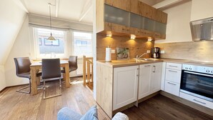 A modern kitchen with wooden cabinets, a built-in oven, and a dining area with a table and chairs. - Vacation apartment 07 south facing with sea view - P: Zollhaus Klein Zicker - exclusive (Thiessow)