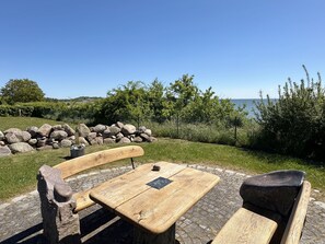 An outdoor wooden table and benches with a stone base, surrounded by a stone wall and greenery, overlooking a body of water.