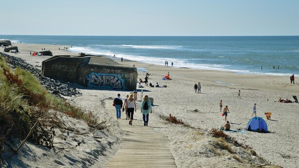 Ein Strand mit einer Holzpromenade, die zum Wasser führt, einer großen, mit Graffiti bespritzten Struktur und Menschen, die den Strand genießen.