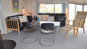 Ein gemütliches Wohnzimmer mit einer Couch, zwei Sesseln, einem Couchtisch und einem Esstisch. Kerzen stehen auf dem Tisch, ein Gemälde an der Wand und ein Fenster mit Vorhängen.