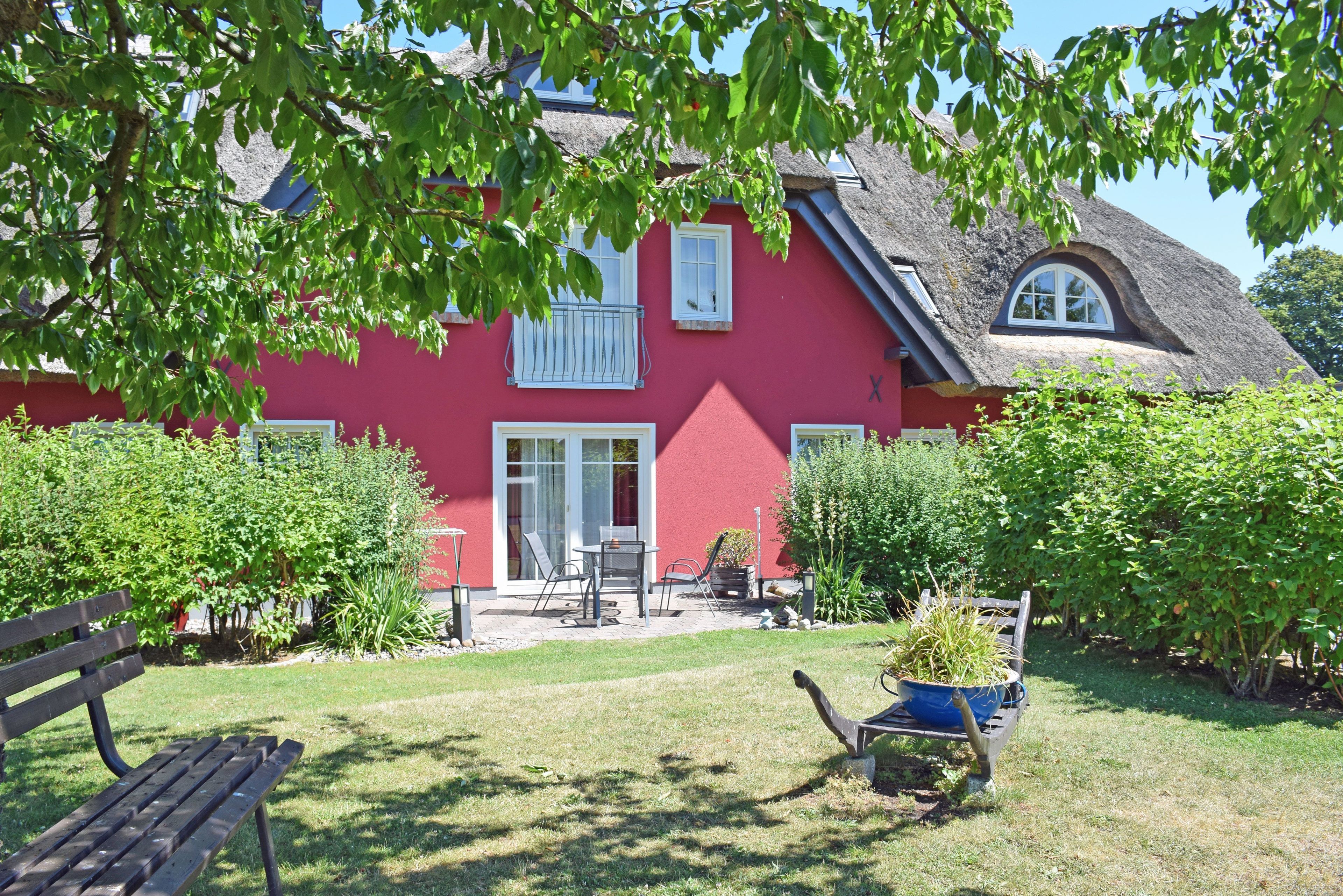 A red house with a thatched roof, a garden with a bench, and a wheelbarrow with plants.