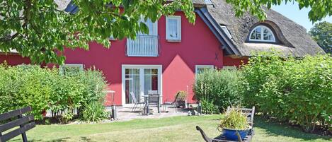 A red house with a thatched roof, a garden with a bench, and a wheelbarrow with plants.