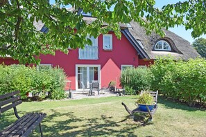 Een rode woning met rieten dak, een tuin met een bankje en een kruiwagen vol planten.