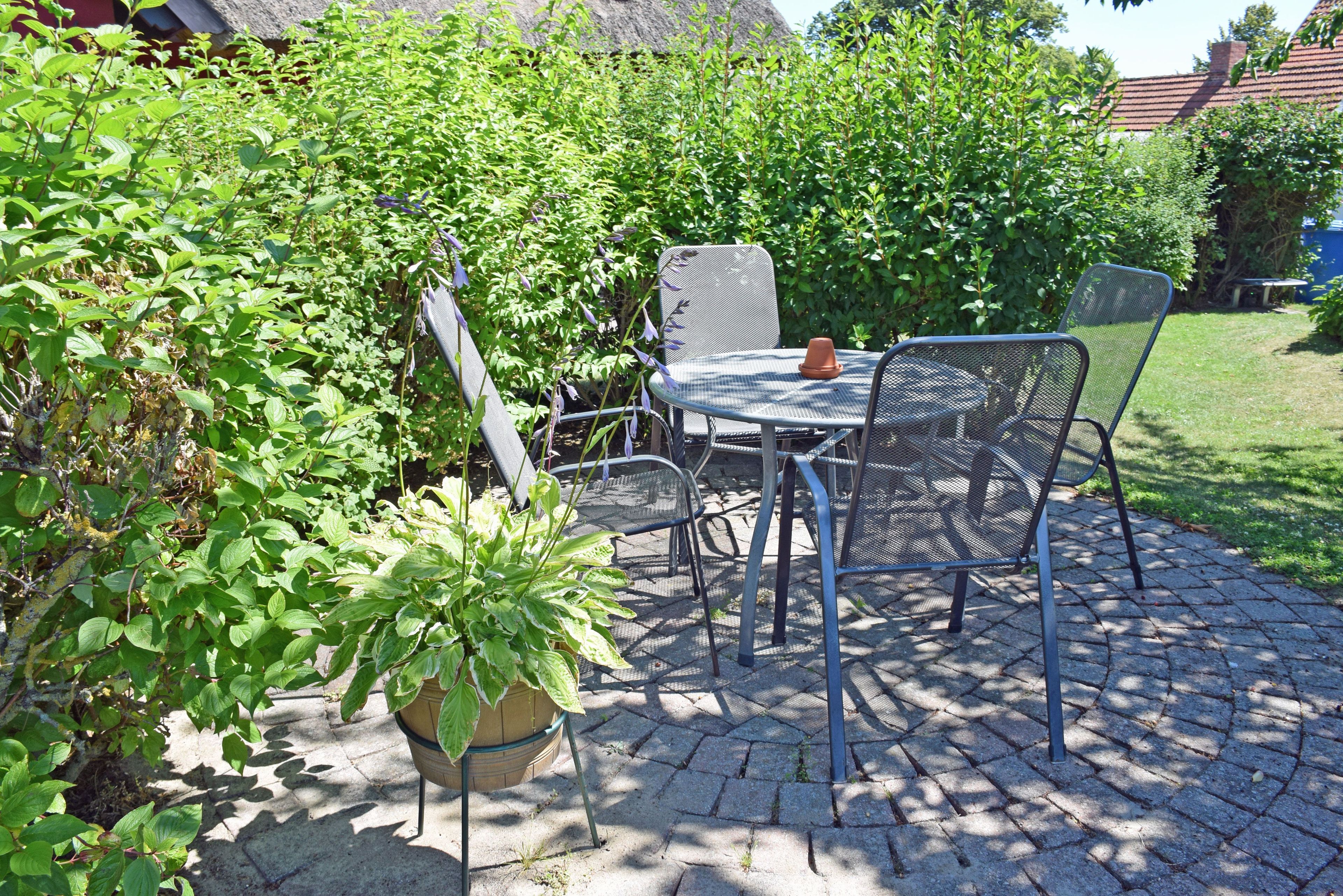 A patio with a round metal table and chairs, surrounded by greenery and a thatched-roof building.