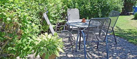 Un patio con una mesa y sillas de metal redondas, rodeado de vegetación y un edificio con techo de paja.