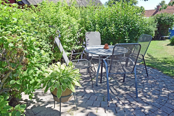 A patio with a round metal table and chairs, surrounded by greenery and a thatched-roof building. - 04 Vacation apartment with lake view - Karolas Landhus under the thatched roof (Mönchgut)