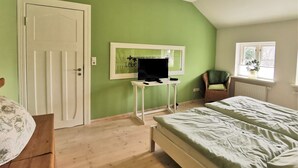 Ett sovrum med en grön vägg, en säng med ett mönstrat överkast, ett skrivbord med en dator, en stol och ett fönster med en växt.