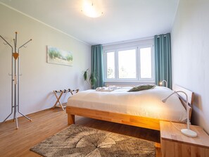 A bedroom with a wooden bed, a bedside table with a lamp, a standing heater, a coat rack, a painting on the wall, and a window with curtains. - Dune garden Whg. Wa45-41 - Wa45-41 (Kühlungsborn)