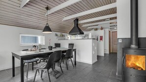 A modern kitchen with a central island, black pendant lights, a wood-burning stove, and a dining area with chairs and a table.