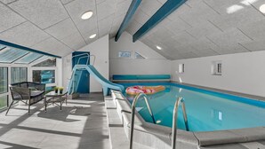 An indoor swimming pool with a slide, a lounge area with chairs, and a view of the outdoors through large windows.