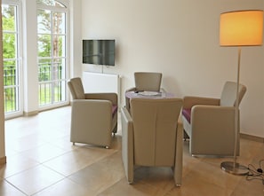 A room with a dining table, four chairs, a television on the wall, and a floor lamp. - Haus Meeresblick FeWo Regenbogen A 1.19 Ref. 132408 u 1 - FeWo Regenbogen A 1.19 (Baabe)