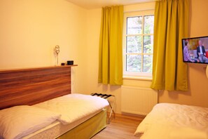Ein Zimmer mit zwei Betten, einem Fenster mit Vorhängen, einem Heizkörper und einem an der Wand befestigten Fernseher.