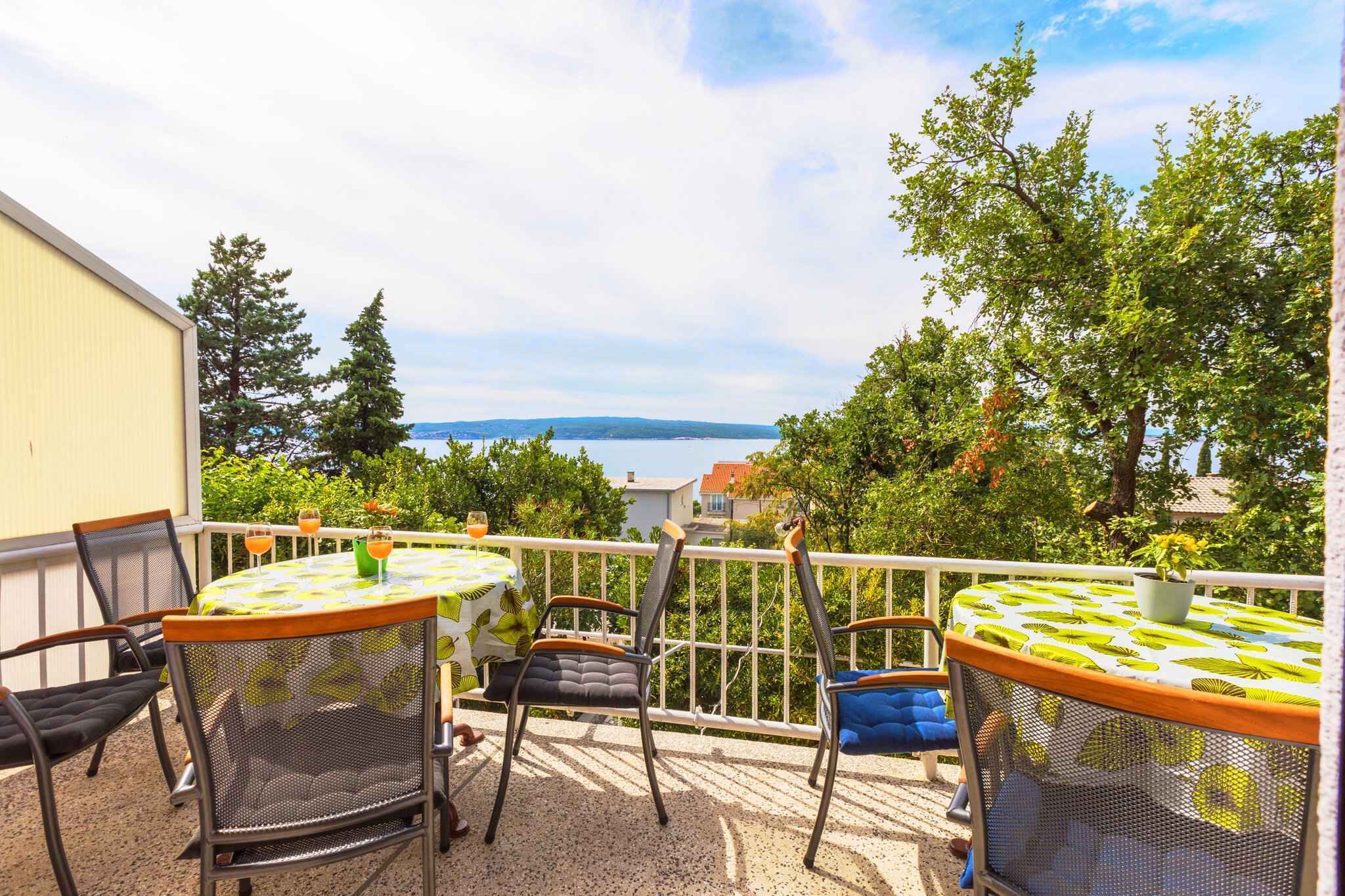 A balcony with a table and chairs, a potted plant, and a view of the sea and hills.